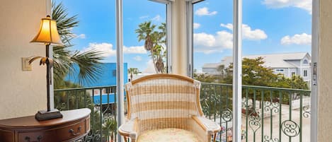 A balcony with a wicker chair, a small table with a lamp, and a view of the ocean and buildings.