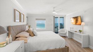 A modern bedroom with a large bed, a view of the ocean, a ceiling fan, and a small desk with a lamp. - Stunning Condo at Crystal Sands on Siesta Key! (Siesta Key)