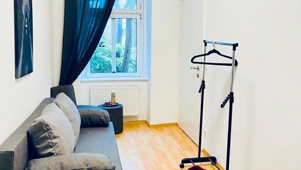 A room with a grey sofa, a window with curtains, a radiator, and a clothing rack. - Comfortable Room by Währinger Park, Shared Apt (Vienna)