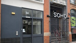 A building with a large glass door marked with the number 34, a metal gate with the word 'SOHO' above it, and a staircase leading to an upper level.