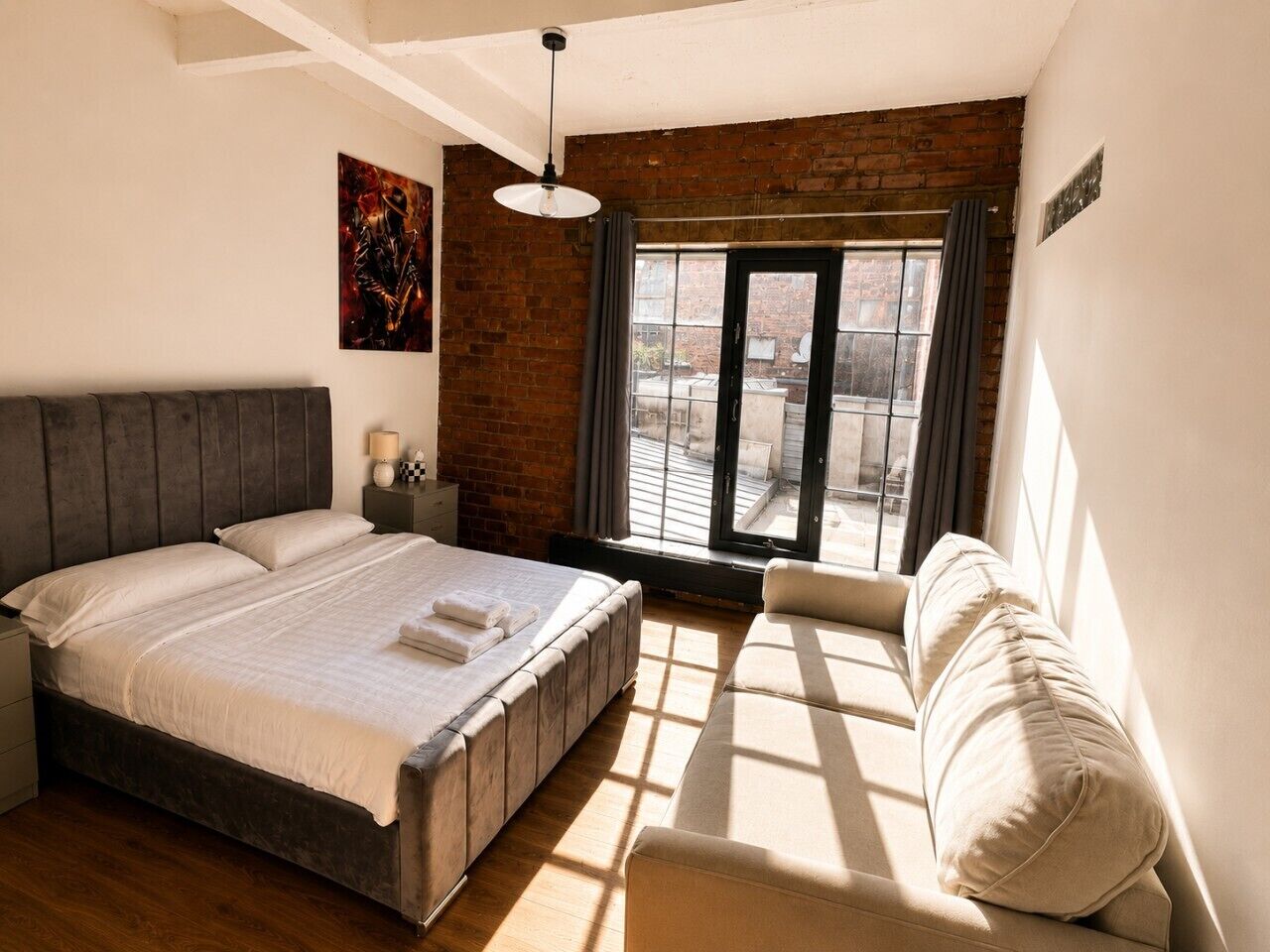 Un dormitorio con cama, sofá, ventana con cortinas, pared de ladrillo y lámpara colgante.