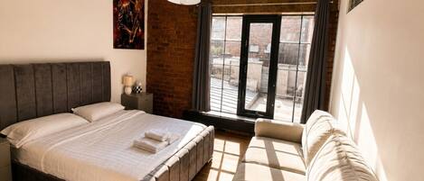 Un dormitorio con cama, sofá, ventana con cortinas, pared de ladrillo y lámpara colgante.