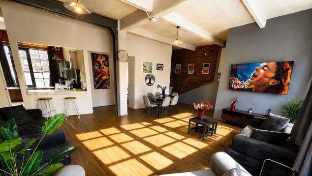 A spacious living area with a large window, a dining table, and a TV screen displaying an image.