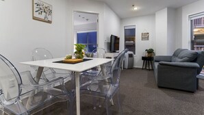 Una sala de estar moderna con una mesa blanca, sillas transparentes, un sofá gris y un televisor.