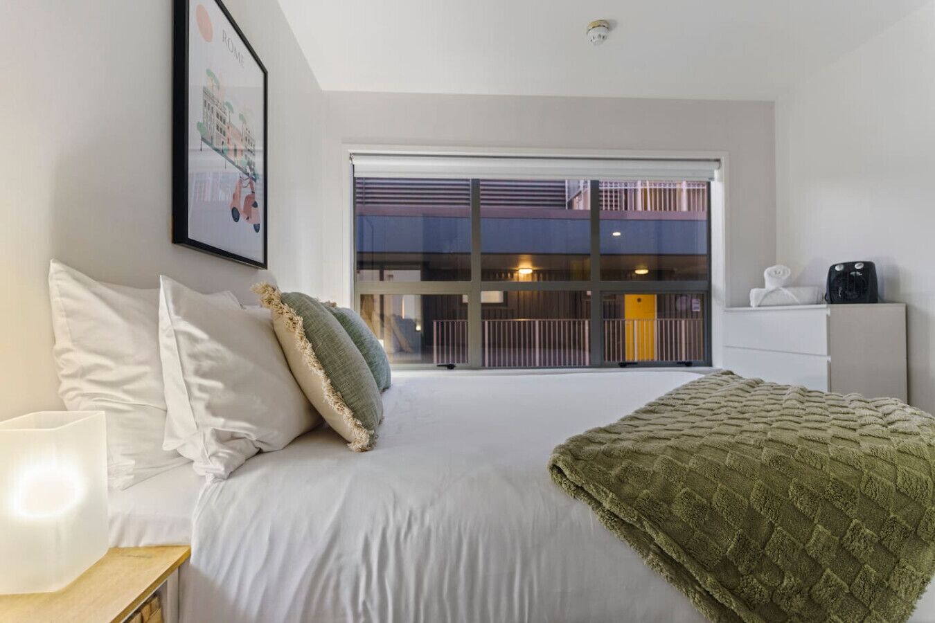 A hotel room with a bed, white pillows, a green throw blanket, a bedside table with a lamp, and a large window with a view of a building and a balcony.