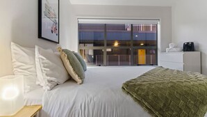 Habitación de hotel con cama, almohadas blancas, manta verde, mesita de noche con lámpara y un amplio ventanal con vista a un edificio y un balcón.