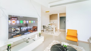 Una sala moderna con un televisor de pantalla plana mostrando una interfaz de YouTube, un comedor con una mesa y sillas blancas, y un sillón naranja brillante.