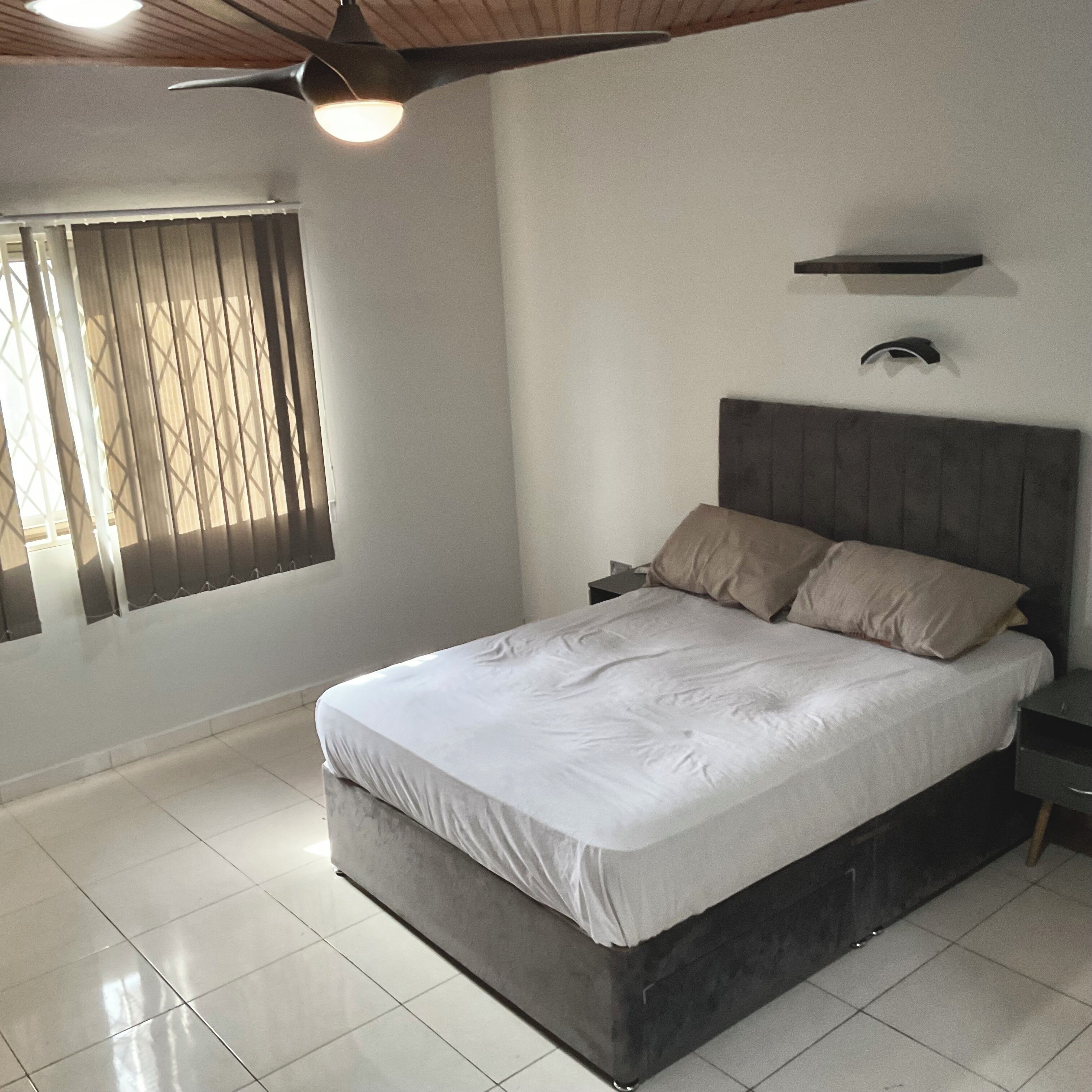 A bedroom with a bed, a nightstand, a ceiling fan, a window with blinds, and a wall-mounted light fixture.