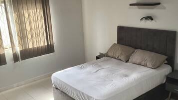 A bedroom with a bed, a nightstand, a ceiling fan, a window with blinds, and a wall-mounted light fixture.