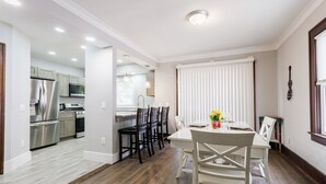 Een moderne keuken met roestvrijstalen apparatuur, een eethoek met een witte tafel en stoelen, en een groot raam met jaloezieën.