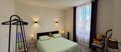 A hotel room with a bed, bedside lamps, a hanging chair, a teddy bear, and a window with curtains.