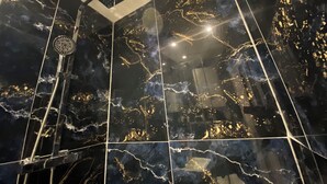Un baño moderno con baldosas de mármol negro y detalles dorados, que incluye una ducha con rociador cuadrado y un cabezal de ducha manual.