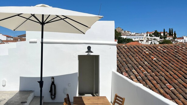 A rooftop terrace with a white umbrella, a wooden table, and a view of terracotta roof tiles and white buildings in the background.