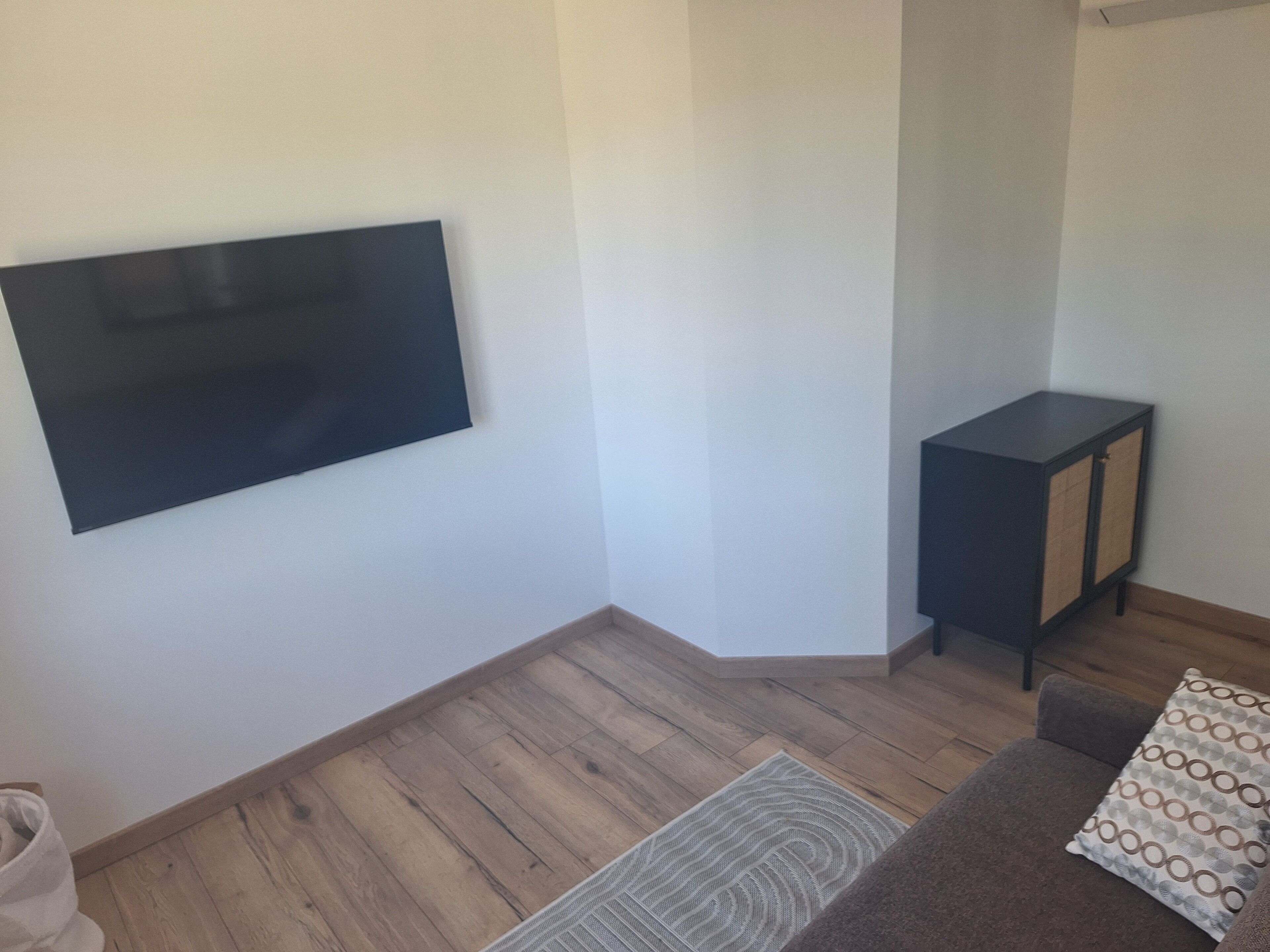 Una habitación con una televisión de pantalla plana montada en la pared, un pequeño armario negro, un sofá marrón con un cojín estampado y suelo de madera.