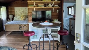 A cozy cabin interior with a wooden ceiling, a bed with a view, a dining area with stools, and a kitchen with cabinets.