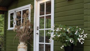 A green wooden house with a white door and windows. There is a vase with dried plants and a potted plant with white flowers on the porch.