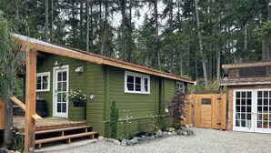 A green wooden cabin with a porch, a wooden deck, and a gravel driveway.