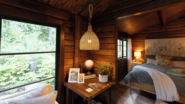 A cozy cabin interior with a wooden ceiling, a desk with a globe and a plant, a bed with a floral headboard, and a large window overlooking greenery.