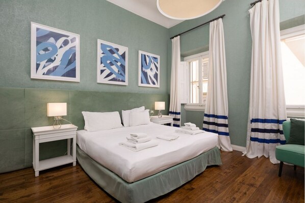 A neatly made bed with white linens and folded towels, a bedside table with a lamp, and a window with curtains.