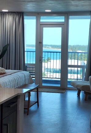 Une chambre avec un lit, une chaise, une table et une vue sur l’océan, visible à travers une porte-fenêtre coulissante.