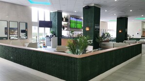 Een moderne receptie met een groen gestructureerd aanrecht, zitplaatsen en een bar met een televisie waarop een sportwedstrijd wordt getoond.
