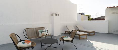 Una terraza en la azotea con sillas de mimbre y una pequeña mesa redonda, paredes blancas y un cielo despejado.