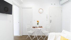 Ein modernes, minimalistisches Wohnzimmer mit einem weißen Sofa, einem kleinen Esstisch mit zwei Stühlen und einer Uhr an der Wand.