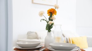 Ein Esstisch mit weißen Tellern, Schüsseln und Besteck, dazu eine Vase mit Blumen und eine Kerze.