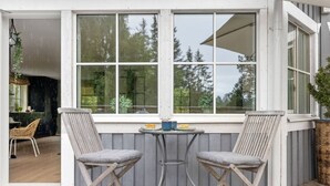 Un espacio de estar al aire libre techado con dos sillas y una mesita, una ventana con vista a árboles y una puerta que da a un espacio interior.