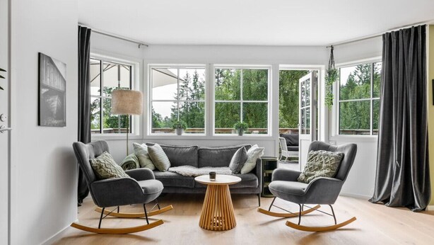Una sala de estar moderna con un sofá gris, dos mecedoras, una mesa de centro redonda de madera y ventanas amplias que dan a zonas verdes.