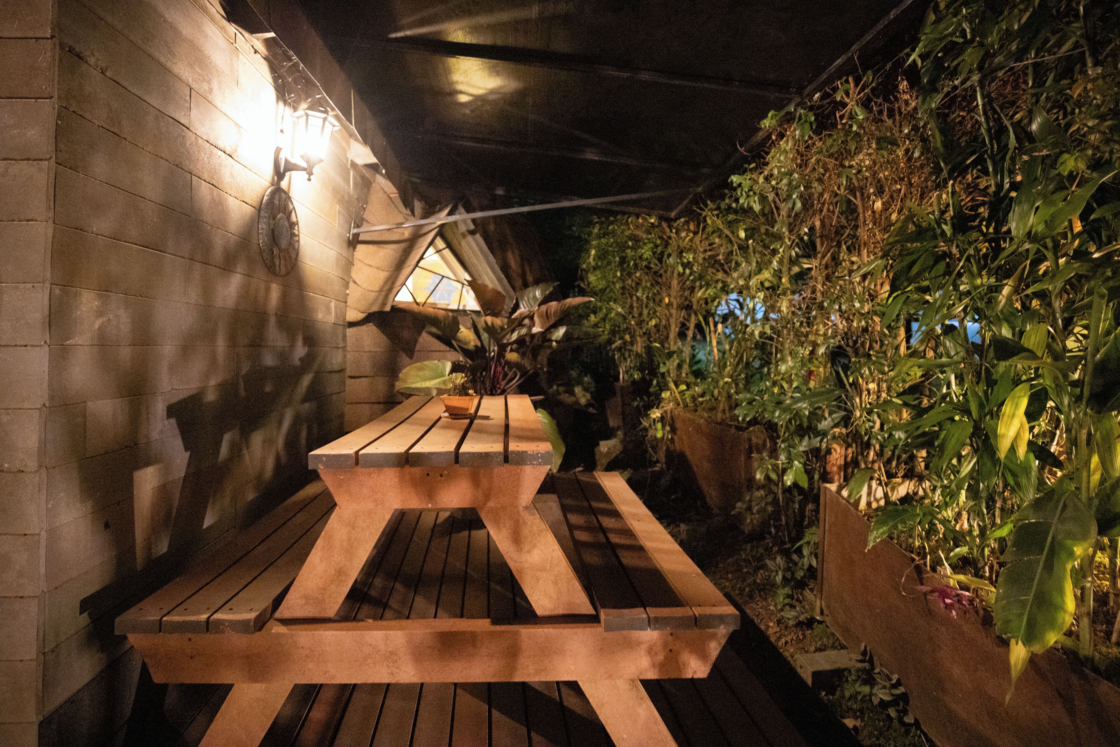 A wooden bench under a sloped roof with a wall of greenery on one side and a brick wall on the other.