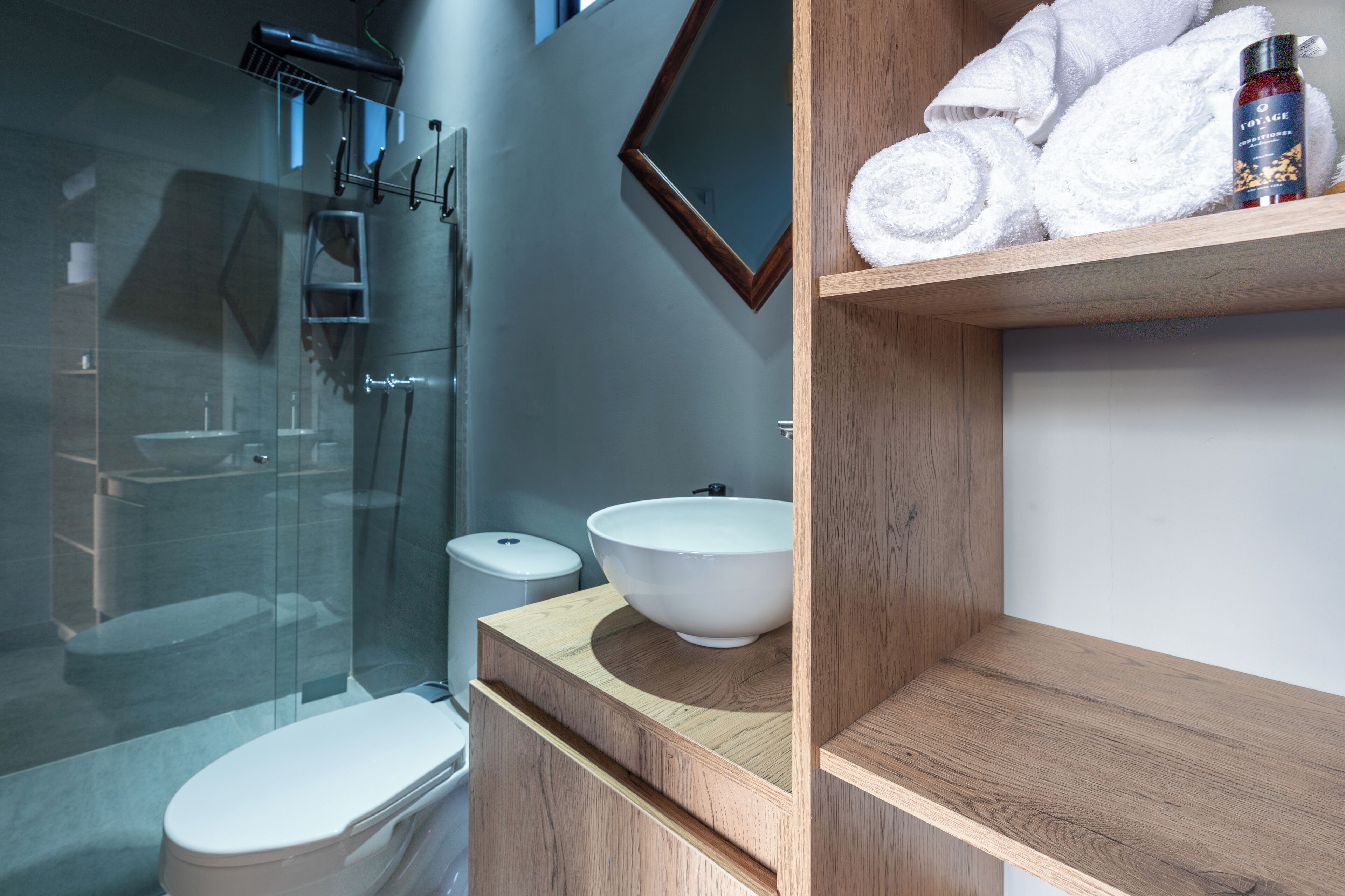 Un baño moderno con cabina de ducha de vidrio, inodoro blanco, tocador de madera con lavabo y estante con toallas enrolladas y artículos de aseo.