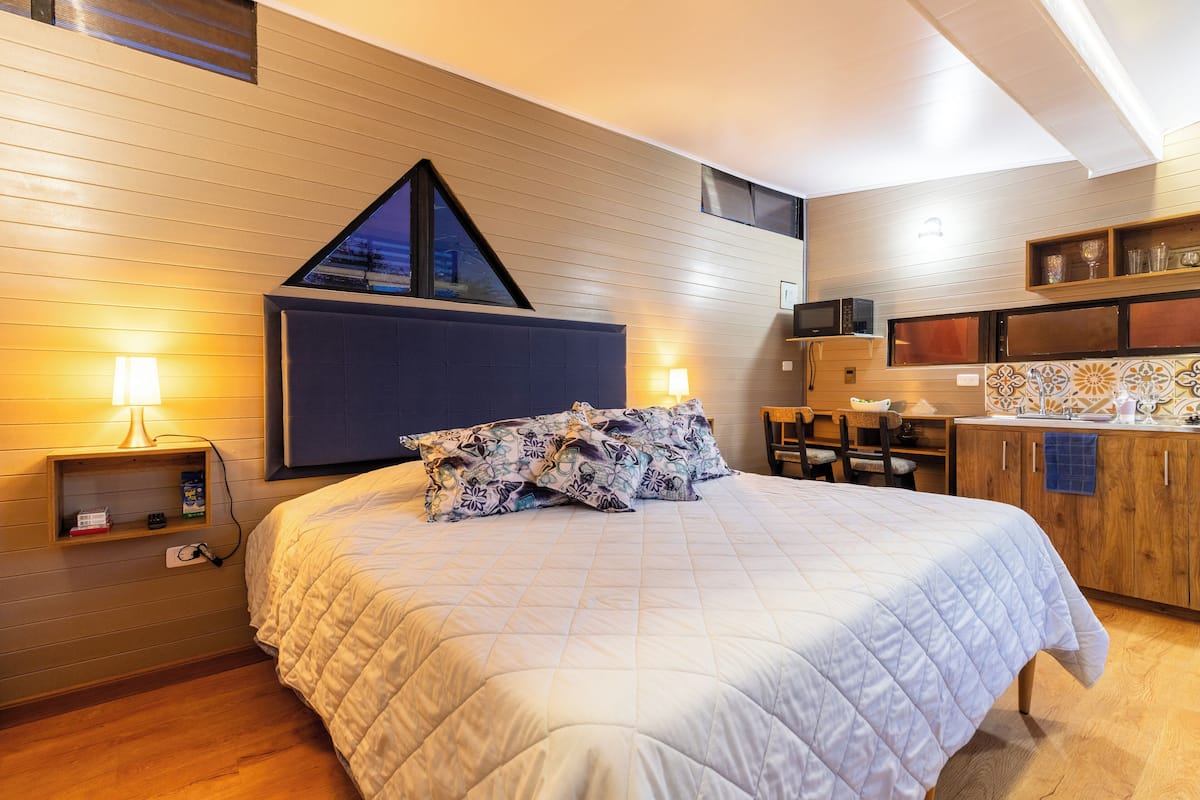 A bedroom with a large bed, bedside lamps, a wooden headboard, a small desk, and a kitchenette area.