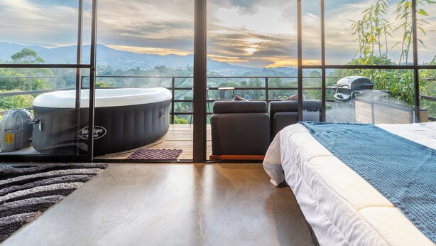A modern bedroom with a large bed, a hot tub, and a view of mountains and greenery. - Roots Eco House (El Peñol)