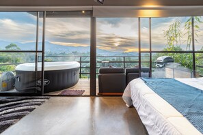 A modern bedroom with a large bed, a hot tub, and a view of mountains and greenery.