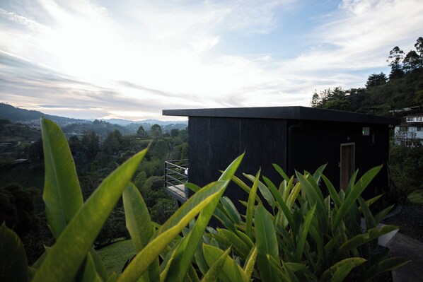 A modern building with a dark exterior, surrounded by lush greenery and a view of distant hills.