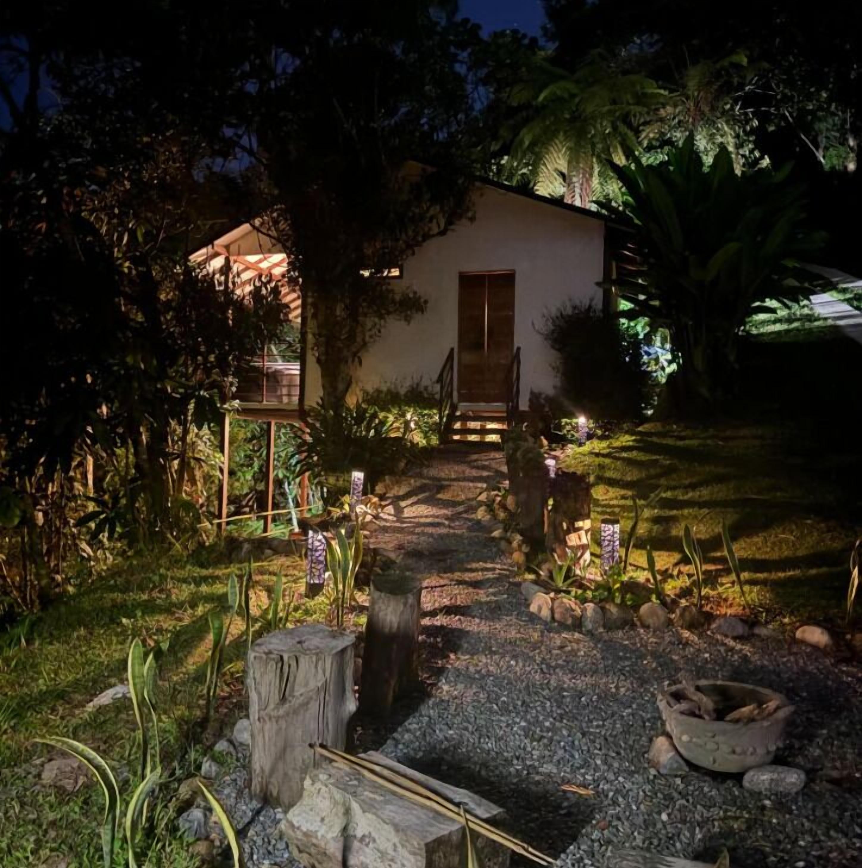 A small house with a wooden door and a porch, surrounded by a garden with various plants and a gravel path leading to the door.
