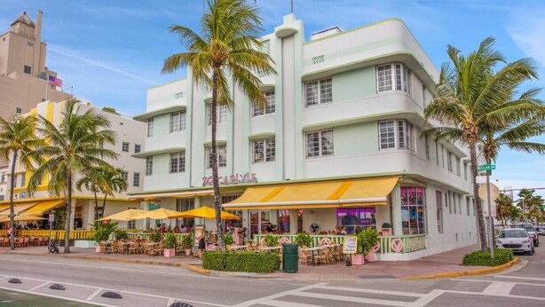 A multi-story building with a green facade, outdoor seating with yellow umbrellas, and palm trees.