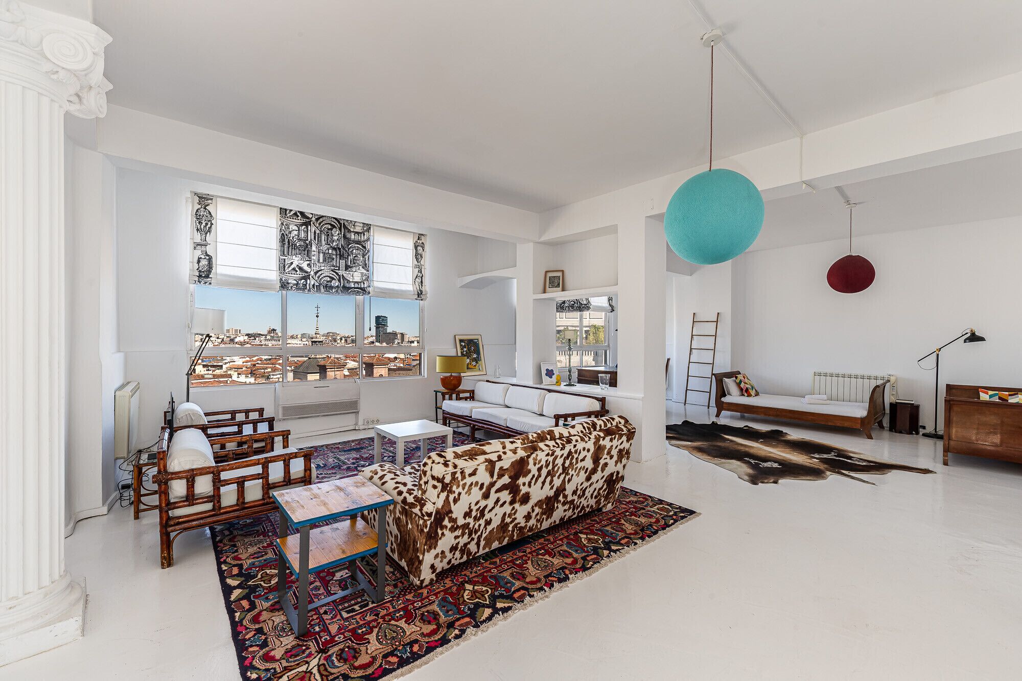 A spacious living room with a large window offering a city view, a patterned sofa, a wooden coffee table, and a hanging blue pendant light.
