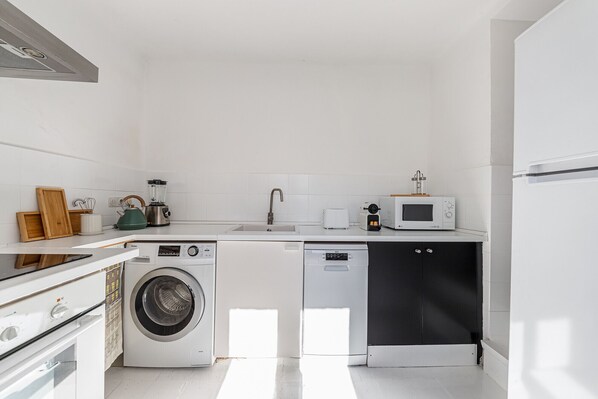 Une cuisine compacte équipée d’un lave-linge, d’un lave-vaisselle, d’un four à micro-ondes et d’un petit plan de travail avec une bouilloire et un grille-pain.