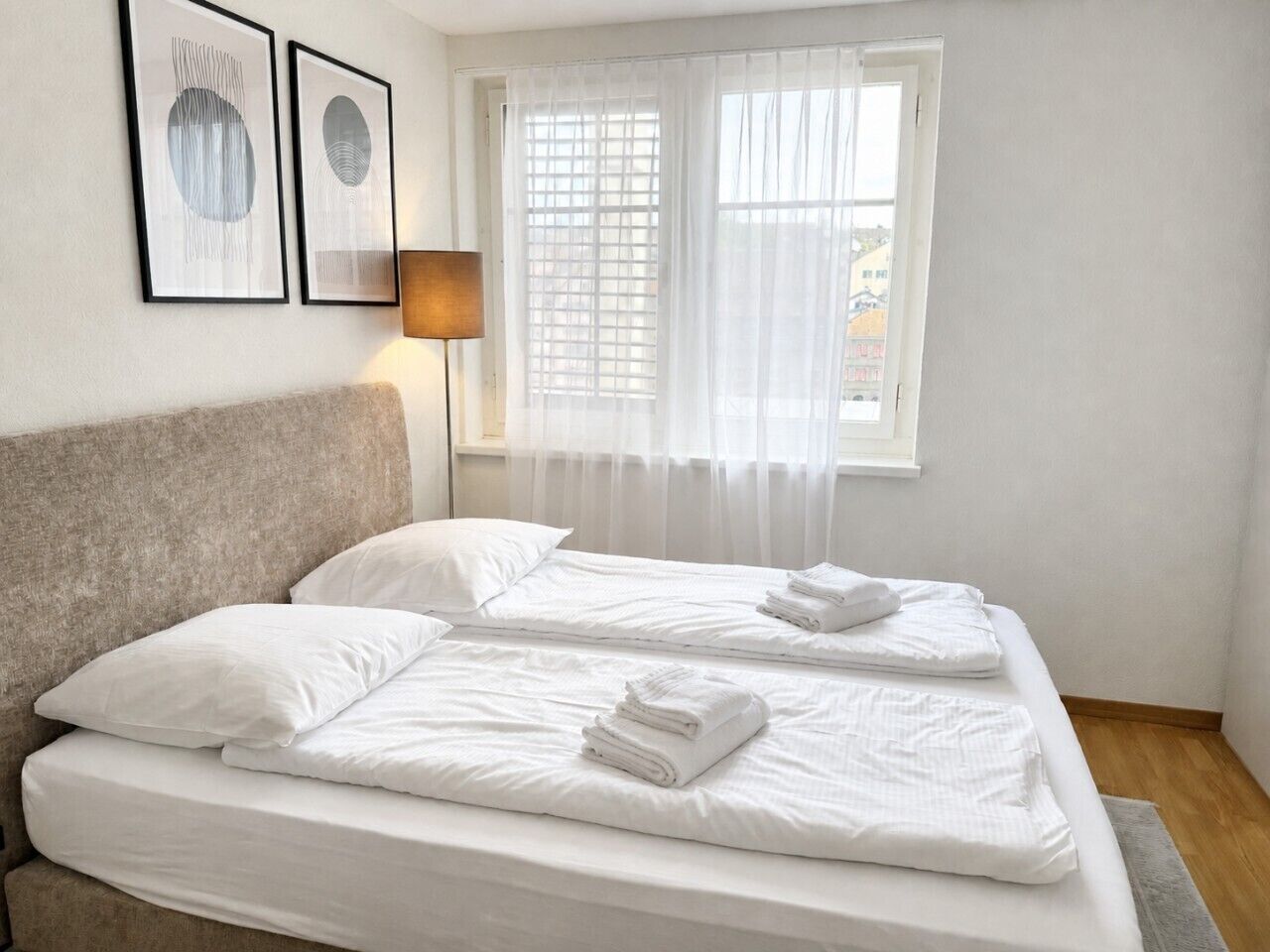 Un lit double avec du linge de lit et des serviettes blanches, une tête de lit, une lampe de chevet et deux œuvres d’art abstraites encadrées accrochées au mur.