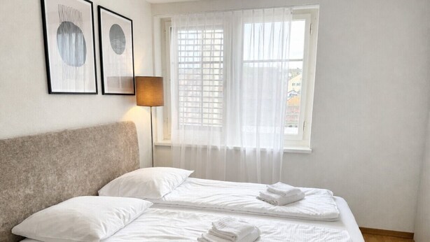 Un lit double avec du linge de lit et des serviettes blanches, une tête de lit, une lampe de chevet et deux œuvres d’art abstraites encadrées accrochées au mur.
