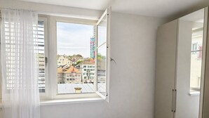 A bedroom with a bed, a window with a view of buildings, and a wardrobe. - Charming Riverside Studio - Stork 41 (Zürich)