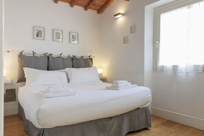 Una cama bien tendida con sábanas blancas y ropa de cama gris, techo de madera y una ventana con cortinas translúcidas.