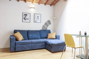 Un sofá azul en forma de L con dos cojines, una silla amarilla y una mesa de vidrio con dos plantas en macetas.