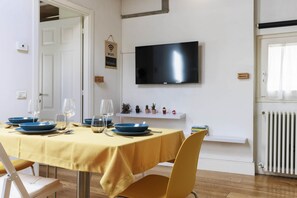 Un comedor con una mesa dispuesta para cuatro, un televisor de pantalla plana en la pared y un estante con pequeños objetos decorativos.