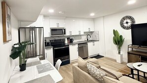 A modern kitchen with white cabinets, a black stove, and a microwave. There is a dining table with chairs and a beige sofa. A plant is on the table, and a clock is on the wall. - Cozy 2 Bedroom Suite Close to Amenities (Edmonton)