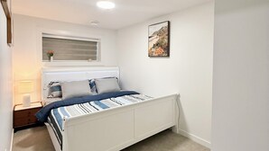 A bedroom with a white bed, a bedside table with a lamp, a window with blinds, and a wall-mounted picture. - Cozy 2 Bedroom Suite Close to Amenities (Edmonton)