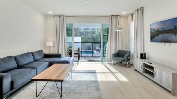 A modern living room with a sofa, coffee table, and TV stand. There is a glass door leading to a balcony with a pool.