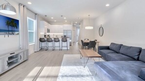 A modern living room with a grey sofa, a wooden coffee table, and a TV stand. There is a kitchen area with a dining table and bar stools.